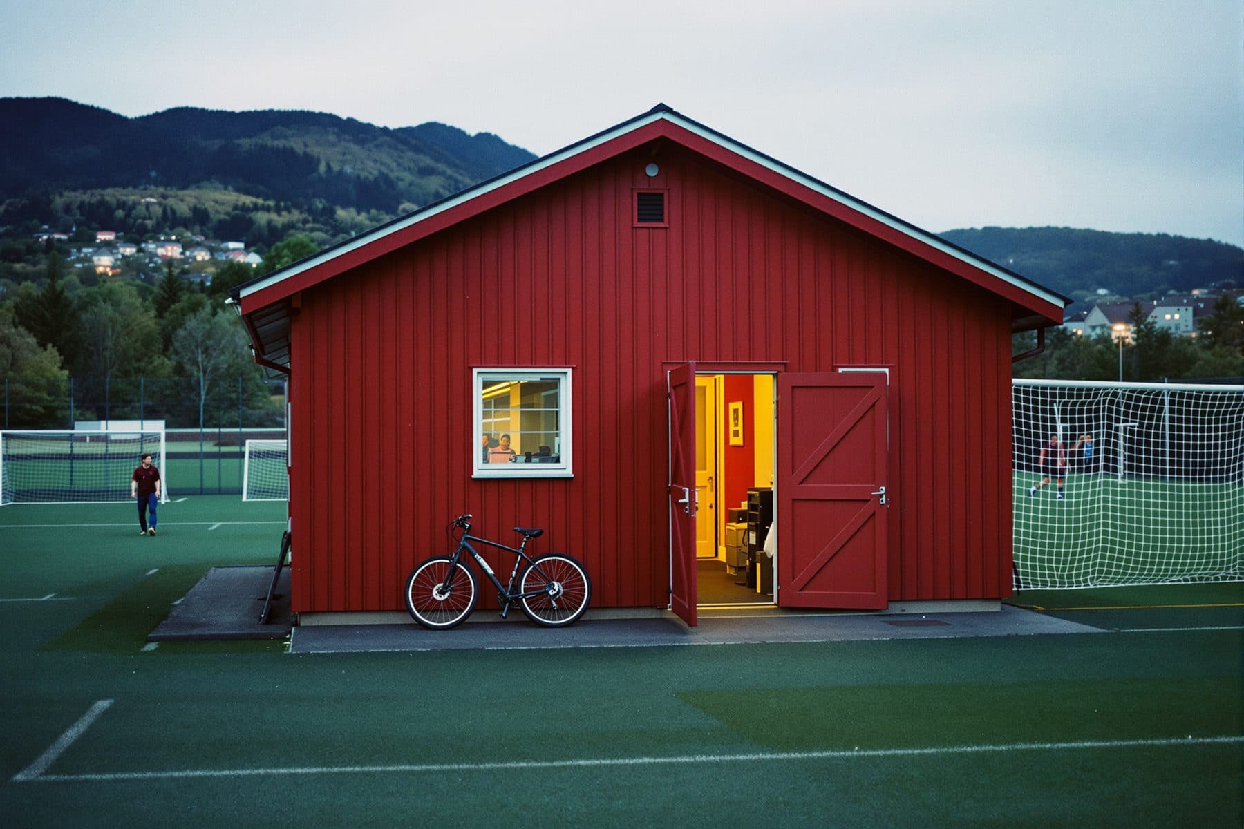 Rødt klubbhus ved en kunstgressbane i kveldslys